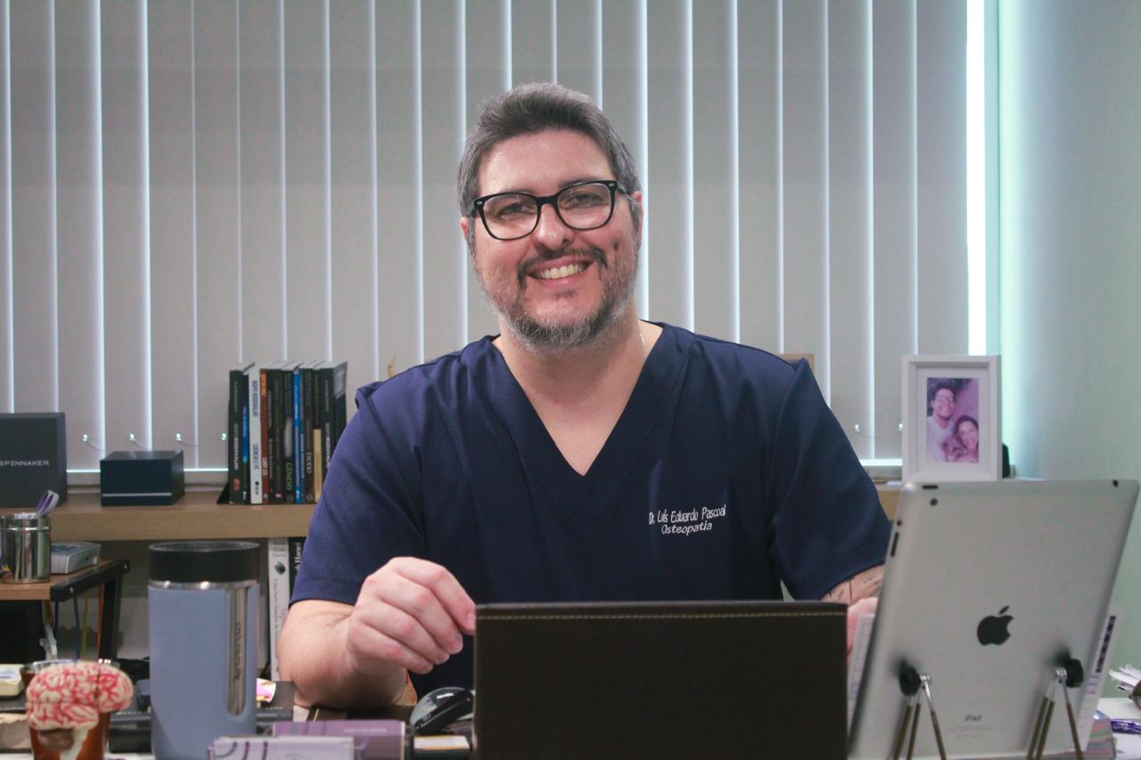 Dr. Luís Eduardo Pascoal, fisioterapeuta e osteopata da EARFF, retrato profissional sorrindo, usando óculos e scrub azul-marinho, sentado em consultório com persianas verticais e estante de livros ao fundo, luz natural suave, fotografia corporativa realista