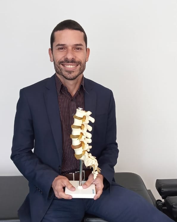 Dr. Angel Mello, fisioterapeuta da EARFF, retrato profissional sorrindo, vestindo terno azul-marinho, sentado segurando modelo anatômico de coluna vertebral, consultório clínico com parede clara ao fundo, luz natural suave, fotografia corporativa realista