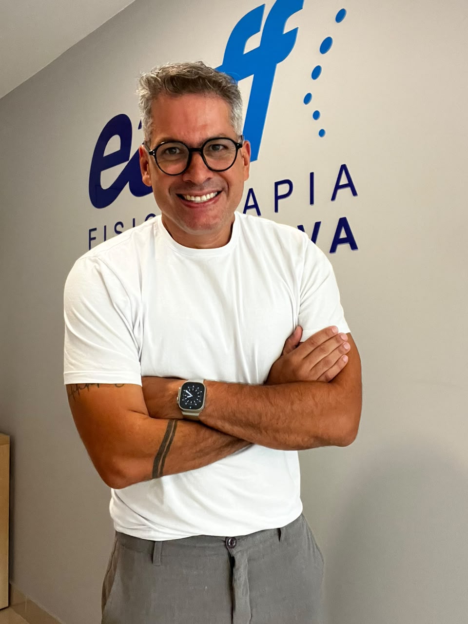 Dr. Fernando Capristano, fisioterapeuta da EARFF, retrato profissional sorrindo com braços cruzados, vestindo camiseta branca, em corredor clínico com logo EARFF ao fundo, luz natural suave, fotografia corporativa