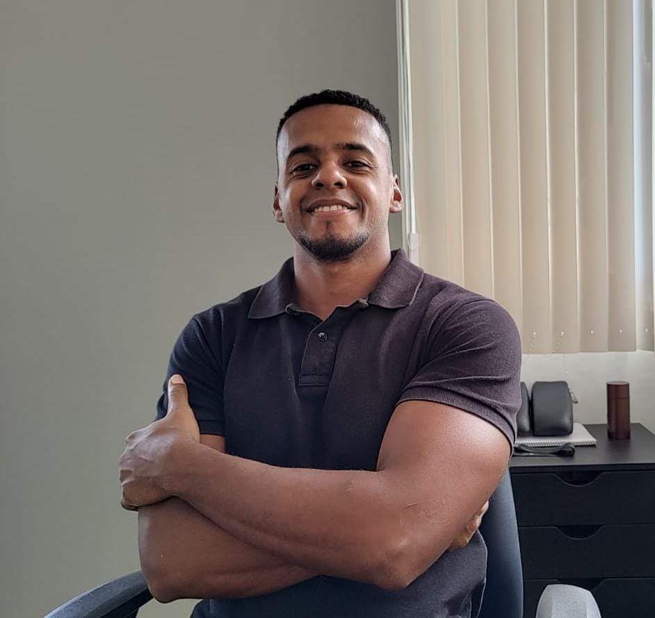 Dr. Raphael Nunes, fisioterapeuta da EARFF, retrato profissional sorrindo com braços cruzados, vestindo polo escura, em consultório clínico com persiana ao fundo, luz natural suave, fotografia corporativa